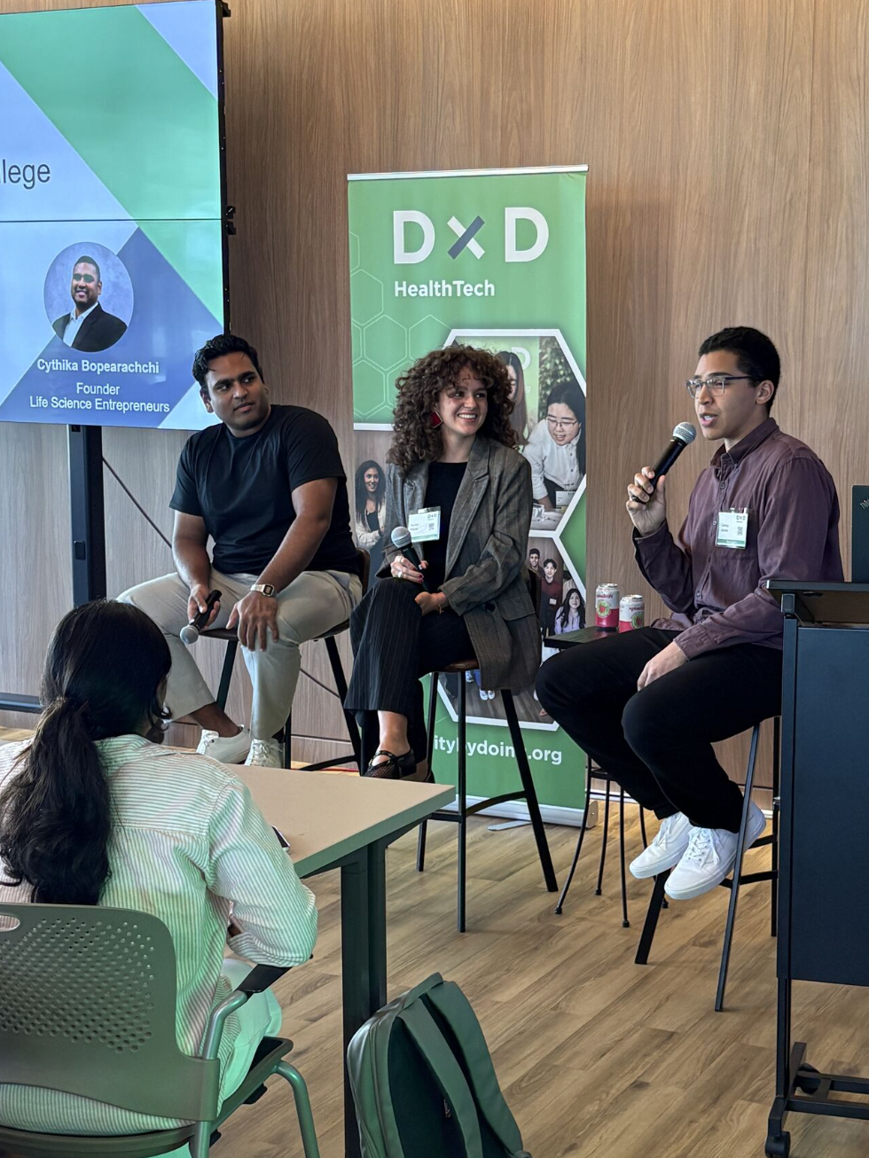 Panel of speakers on stools beside a green DxD HealthTech banner at a HealthTech event