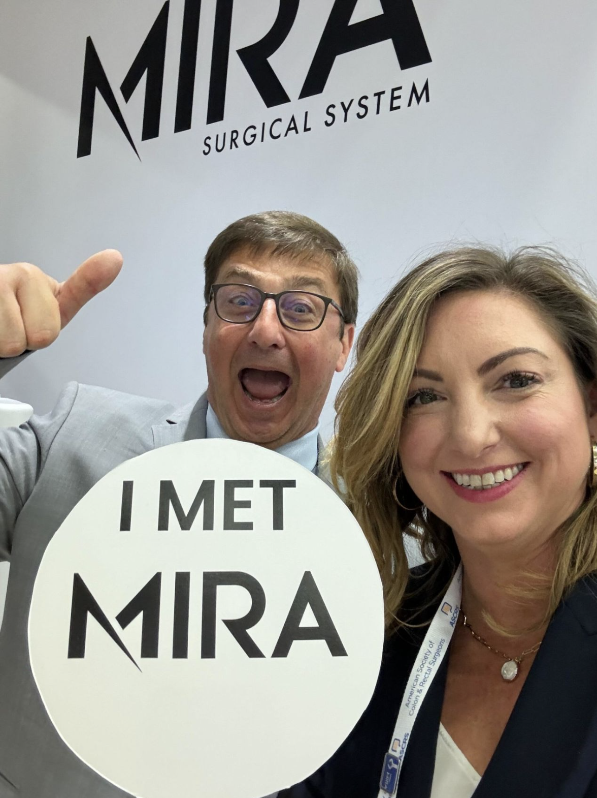 Two attendees holding an I met MIRA sign in front of the MIRA Surgical System backdrop at ASCRS